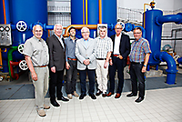 Acht Personen in Zivilkleidung vor blauen Rohrleitungen und Ventilen in einer Wasseraufbereitungsanlage, lächelnd zur Kamera stehend.